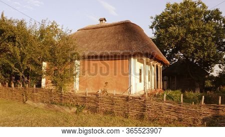 Old East European Rural Wooden House With Wattle Fence And Thatched Roof. Sunny Summer Day.