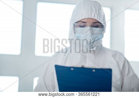 Medical Worker In Protective Clothing, Medical Mask And Goggles Looking At Clip Folder