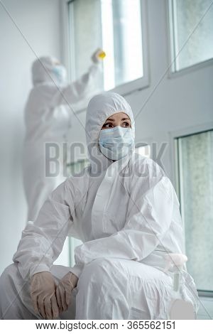 Medical Worker In Protective Clothing And Medical Mask Sitting Next To The Window, Her Collegue Desi