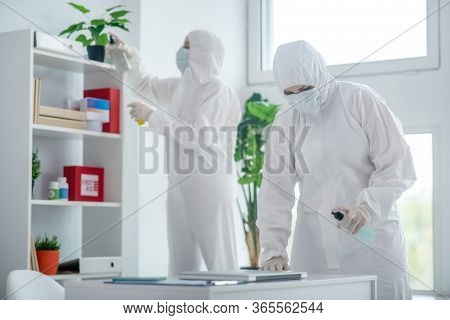 Medical Worker In Protective Clothing And Medical Mask Disinfecting Table, Another Worker Standing B