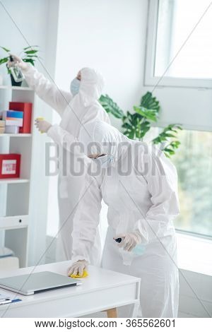 Medical Worker In Protective Clothing And Medical Mask Disinfecting Table, Another Worker Cleaning C
