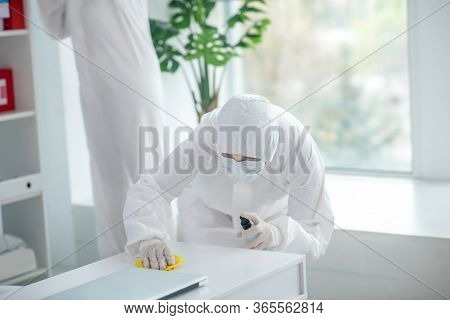 Medical Worker In Protective Clothing And Medical Mask Disinfecting Table, Another Worker Standing B