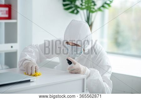 Medical Worker In Protective Clothing And Medical Mask Sprays Disinfectant On Table