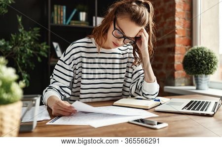 Pensive Diligent Female Freelancer In Glasses And Casual Clothes Analyzing Information In Documents 