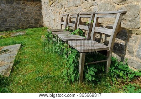 4 Old Wooden Chairs In Overgrowing Grass And Ivy In Front Of A Stone Church Wal.l