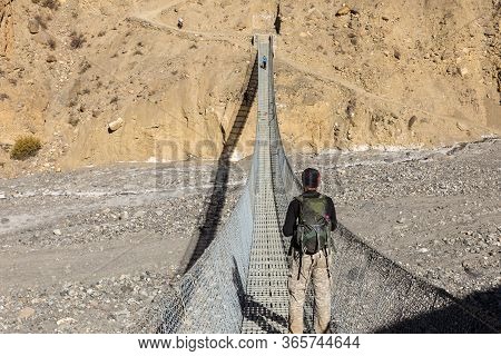 A Man With A Backpack Stands On A Suspension Pedestrian Bridge Over A Mountain Gorge And Looks At Th