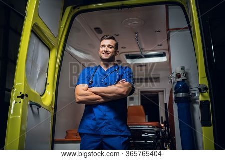 Young Paramedic In A Blue Uniform Standing And Smiling In Front Of An An Ambulance Car