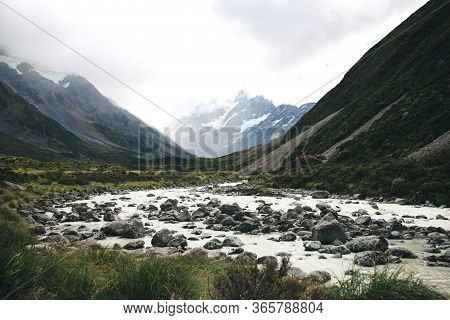 Beautiful Mount Cook In New Zealand With A Cloudy Sky And Green Grasss With A River And Rocks
