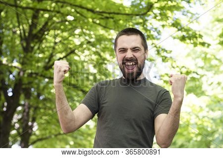 A Bearded Young Caucasian Man, Overjoyed, Walks Through The Park Between The Trees, Raising His Hand