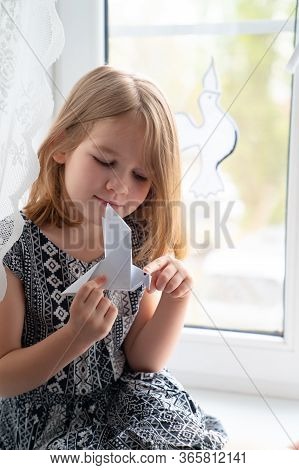 Little Girl Kid Sitting On The Windowsill With Origami In His Hands And On The Window Of Doves Symbo