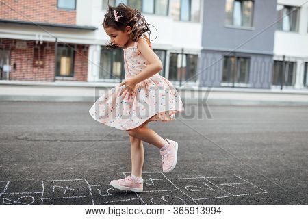Horizontal Image Of Little Girl Playing Hopscotch On Playground Outdoors. Cute Child Plays Next To T