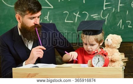 Teacher In Formal Wear And Pupil In Mortarboard In Classroom, Chalkboard On Background. Father Check