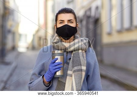 health, safety and pandemic concept - young woman wearing black face protective reusable barrier mask with tumbler or thermo cup on city street
