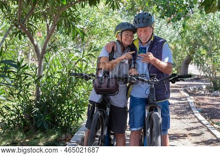 Active Caucasian Senior Couple With Electrobikes Standing Outdoors In The Park Video Calling By Cell