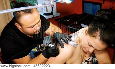 In A Tattoo Salon , A Specialist Is Doing A Tattoo On Womans Back, A Floral Ornament. A Man Works In