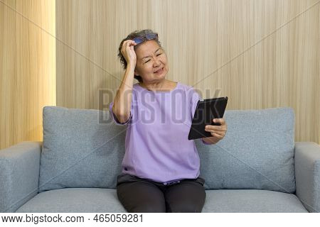 Asian Retired Elderly With Eyeglasses, Sitting On The Sofa Watching At Tablet Computer. With Presbyo