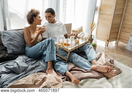 Full Length Of Barefoot African American Couple In Jeans Sitting Near Tray With Coffee And Croissant