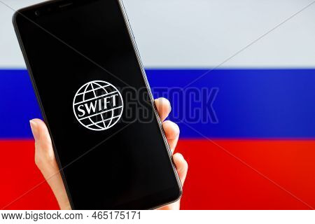 Ukraine, Kiev - February, 23 2022: Hand Holding Mobile With Swift Logo And Russian Flag In Backgroun