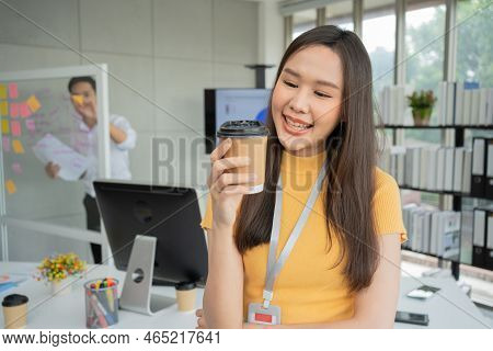 Young Asian Happy Businesswoman Or Intern Holding Coffice Cup In Modern Office Background Is Colleag