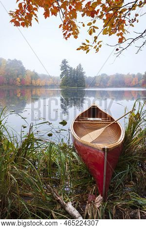 Red Wooden Canoe On Calm Lake With Trees In Fall Color And Maple Branches Above During Autumn
