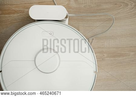 A White Robotic Vacuum Cleaner Stands On The Floor While Charging, Waiting To Be Turned On To Clean 
