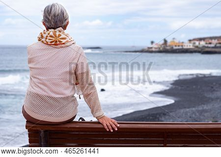 Rear View Of Senior Woman Sitting On Outdoors Bench At Sea Looking At Horizon Over Water. Active Cau