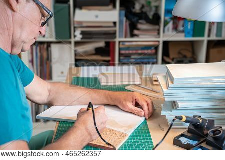Senior Man Working With The Pyrographer Burning The Wood Making Artwork Craft In His Art Studio.