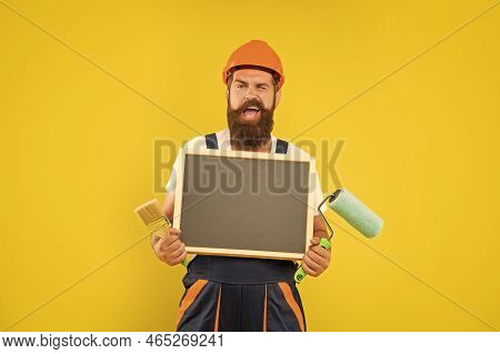 Happy Man Housepainter In Helmet And Work Clothes Hold Paint Roller Brush And Blackboard