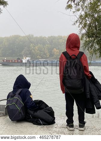 Thoughtful Boys With Hoods Looking A Cago Ship Sailing Down Danube River. Cold And Calm Winter Day C