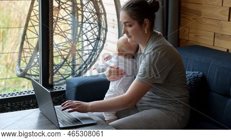 Young Mother Types On Laptop Sitting With Baby Girl On Lap Near Window In Light Room. Little Child T