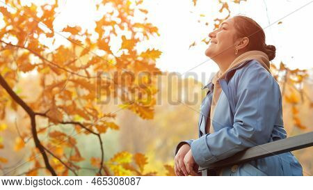 Woman In Blue Coat Stands On Balcony By Park Leaning On Railing With Hands. Female Breathes Deeply A