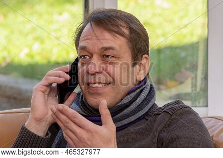 An Elderly Smiling Man Of 45-50 Years Old In A Scarf Is Talking On A Smartphone, Gesturing With His 