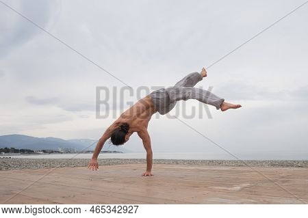 Shirtless Caucasian Man Doing Backflip On Pebble Beach.