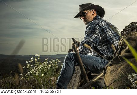 American Cowboy In His 40s Enjoying Free Time On His Countryside Ranch. Seating On A Piece Of Wooden