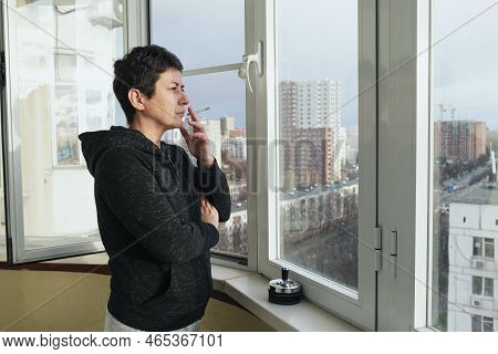 Middle-aged Brunette Woman Stands On The Glazed Balcony Of Her Apartment And Smokes. A Woman With A 