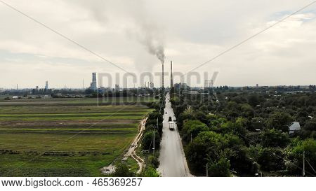 Aero. In Middle Of Field, A Road With Cars That Leads To A Large Plant, Two Tall, Big, Smoking Chimn