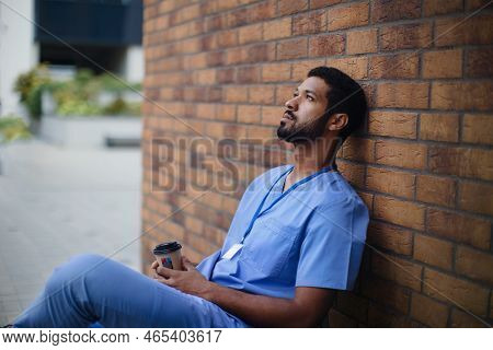 Tired Caregiver Man Resting Outdoor, In Front Of Nurishing Home.
