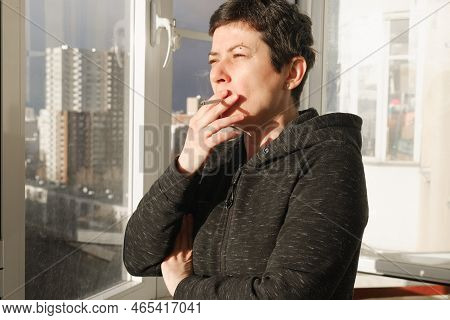 Middle-aged Brunette Woman Stands On The Glazed Balcony Of Her Apartment And Smokes. A Woman With A 
