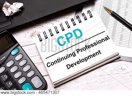 On A Light-colored Office Table Is A Computer, A Black Calculator, A Pen, And A Notebook With The Te