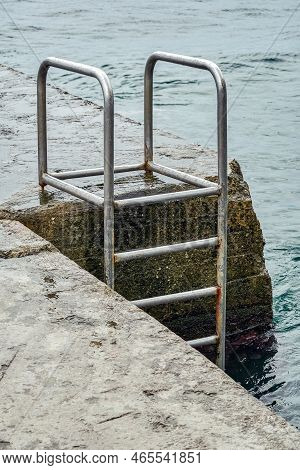 Grey Concrete Pier With Metal Ladder At Blue Sea Water With Small Waves On An Overcast Summer Day