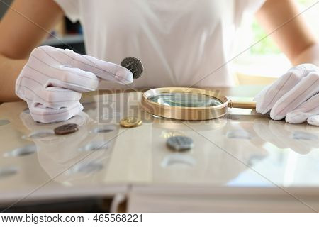 Female Numismatist Examines Collection Of Coins. Woman Looks At Coins Through Magnifying Glass. Numi