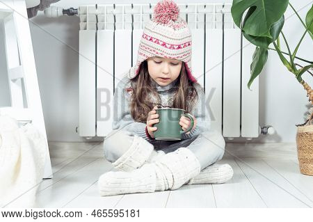 Sad Little Girl In Woolen Hat, Socks And Pullover Drink Hot Tea And Sit With Crossed Legs Near Heati