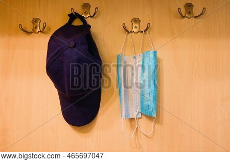 Close-up View Of Coat Rack With Two Medical Masks And Blue Baseball Cap Are Hanging On Wooden Wall. 