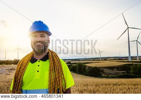 Happy Maitenance Worker Standing In Wind Turbine Farm Field With Climbing Equipment: Helmet And Rope