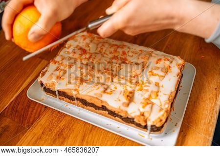 Female Chef Grating Orange Zest Over Walnut Cake With Cream On Wooden Kitchen Table. High Quality Ph