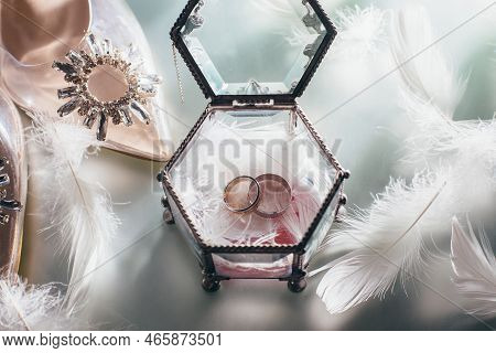 Wedding Gold Rings In A Glass Box With White Feathers Next To The Bride's Shoes Decorated With Stone