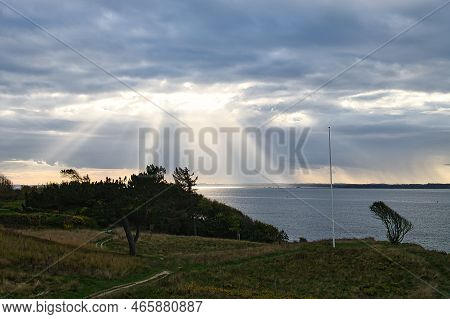 On The Coast Of Hundested. Sun Rays Break Through The Dramatic Sky Through The Clouds. Meadow With P