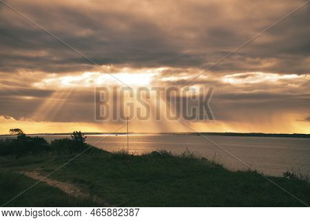 On The Coast Of Hundested. Sun Rays Break Through The Dramatic Sky Through The Clouds. Meadow With P