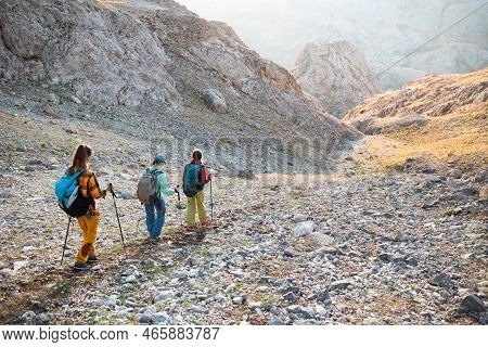 Three Tourists With Backpacks In The Mountains. Tourists Travel Through The Rocky Mountains. Active 