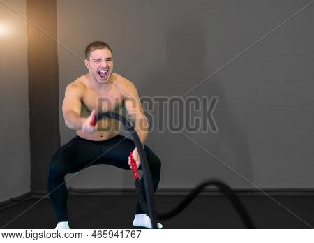 A Handsome Muscular Young Man Using Battle Ropes For Exercise In A Gym. He Focuses On Performing The